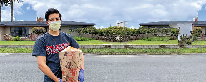 A young man with a mask on standing in front of houses holding a large Trader Joe&apos;s brown bag