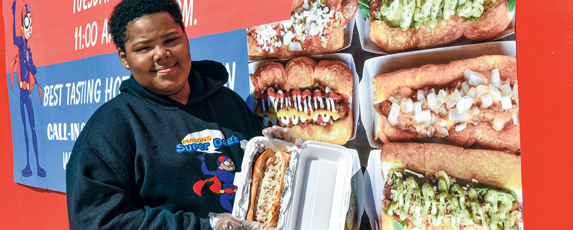 a boy holding a loaded hot dog in front of pictures of other hot dogs