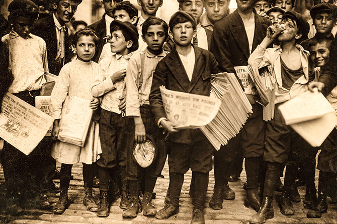 Black and white photo of a group of boys walking and carrying stacks of newspapers
