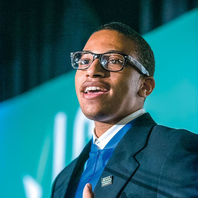 A young man wearing black glasses and a suit talking to an audience
