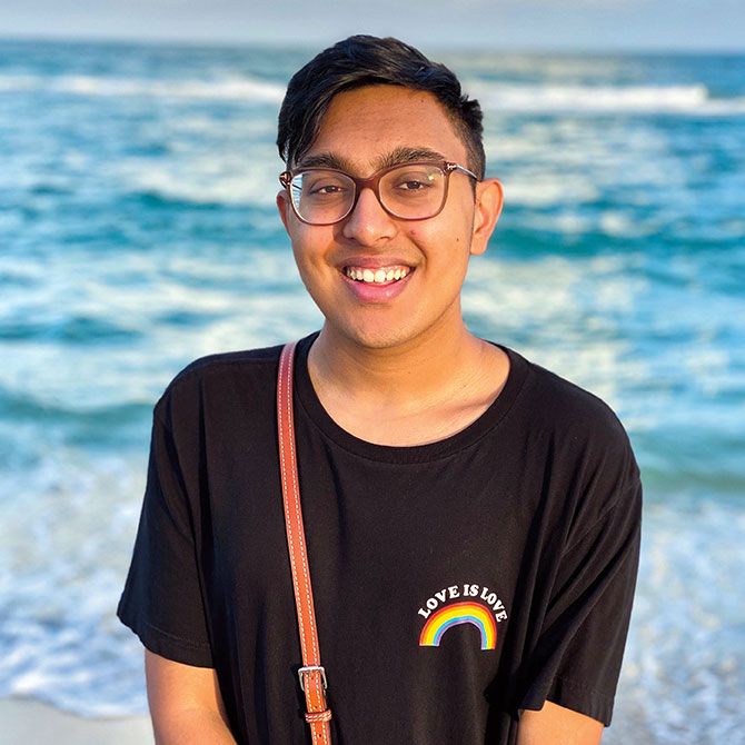 A smiling young man wearing brown glasses and a black shirt with a small raindbow on the chest