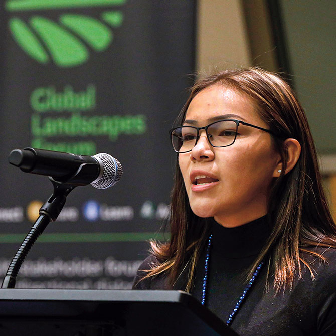A young woman wearing glasses speaking into the podium microphone as she addresses an audience