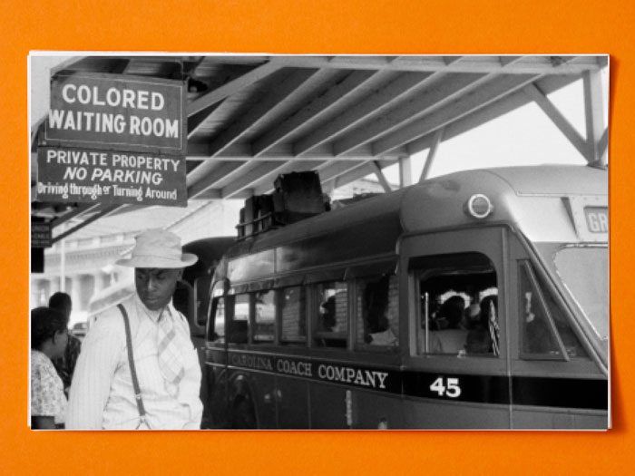 Black and white photo of a man waiting for the train under the sign "Colored Waiting Room"