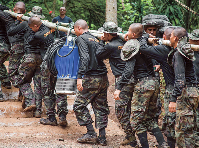 Men in camouflage carrying a long piece of wood with a machine and its wires wrapped around it