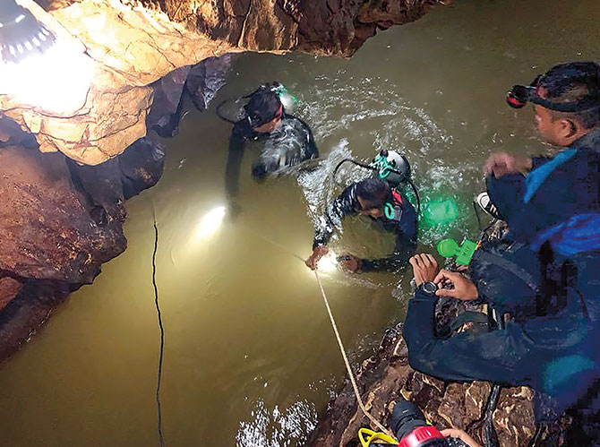 Men with diving gear and a yellow rope standing in water as other men watch from the ground above