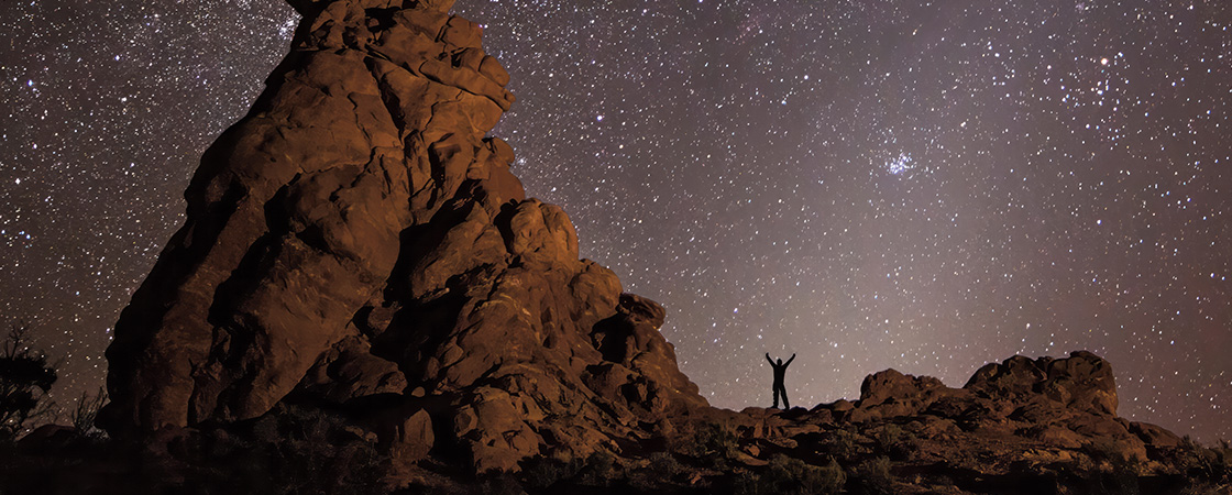 Photo of a person standing next to a desert rock formation at night with endless stars in sky