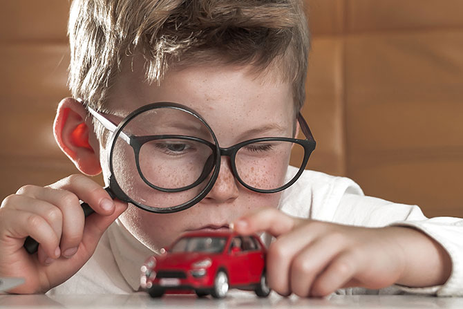 A boy wearing black glasses looking at his toy car through magnifying glass