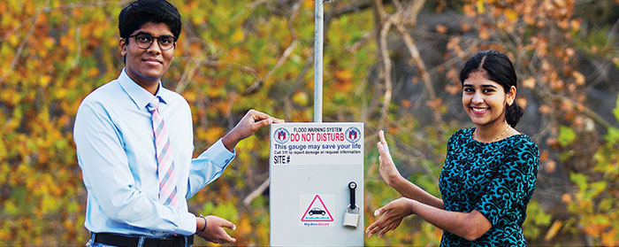 A boy and a girl presenting a metal box  attached to a pole with the text "HydroAlert" on it